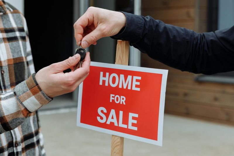 Close-up of a home for sale sign with people exchanging keys